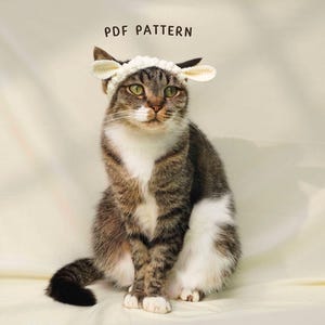 May include: A tabby cat wearing a white crocheted sheep hat with floppy ears. The cat is sitting and looking at the camera. The text "PDF PATTERN" is above the cat.
