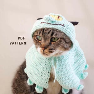 May include: A light mint green crocheted pet hood with a creature-like design featuring a small snout, yellow eyes, and dangling appendages. The hood is worn by a tabby cat and includes the text PDF PATTERN printed on the left side of the image.