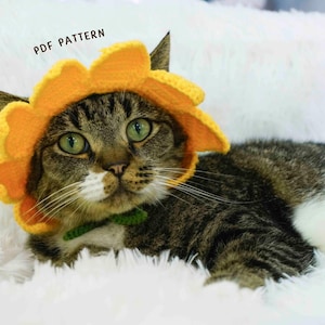 May include: A tabby cat wearing a bright yellow crochet sunflower hat with a green collar. The cat has green eyes and is lying on a fluffy white surface. Text at the top reads "PDF PATTERN".
