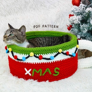 Może przedstawiać: Szydełkowe legowisko dla kota w kształcie świątecznego kubka. Legowisko jest czerwone z zieloną obwódką i białymi akcentami, ozdobione kolorowymi światełkami i napisem "XMAS". W środku siedzi kot.