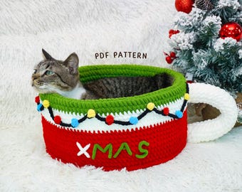 May include: A crocheted cat bed shaped like a Christmas mug. The bed is red with a green rim and white accents, decorated with colorful lights and the word "XMAS". A tabby cat is inside the bed.