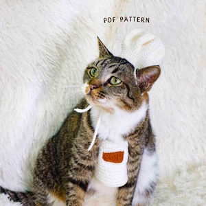 Puede incluir: Un gato atigrado con un gorro de chef y un delantal de ganchillo blancos con un detalle marrón. El gato está sentado sobre una superficie blanca y esponjosa. El texto "PDF PATTERN" es visible.