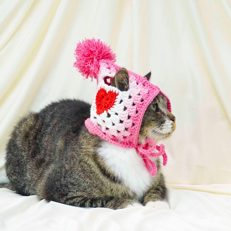 K&ouml;nnte beinhalten: Eine getigerte Katze tr&auml;gt einen rosa-wei&szlig;en geh&auml;kelten Hut mit einem roten Herz und einem rosa Bommel. Der Hut ist mit rosa Garn unter dem Kinn gebunden. Die Katze liegt auf einer wei&szlig;en Oberfl&auml;che.