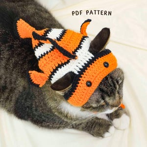 Könnte beinhalten: Eine getigerte Katze trägt einen handgefertigten Häkelhut in Form eines Clownfisches. Der Hut ist orange, schwarz und weiß, mit schwarzen Knopfaugen. Der Text "PDF PATTERN" ist sichtbar.