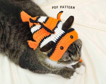 May include: A tabby cat wearing a handmade crochet hat in the shape of a clownfish. The hat is orange, black, and white, with black button eyes. The text "PDF PATTERN" is visible.