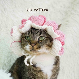 Könnte beinhalten: Eine getigerte Katze trägt einen handgefertigten Häkelblumenhut mit rosa und weißen Blütenblättern. Der Hut ist mit einer weißen Schnur unter dem Kinn gebunden. Der Text "PDF PATTERN" befindet sich über dem Kopf der Katze.
