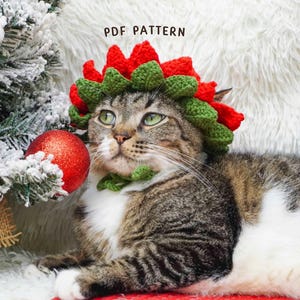 May include: A tabby cat wearing a crocheted Christmas wreath and collar, with red and green accents. The cat is lying down near a decorated Christmas tree and a red ornament. The text "PDF PATTERN" is visible.