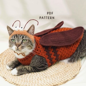 May include: A tabby cat wearing a handmade crochet bee costume, including a hat with antennae and a body suit with wings. The costume is primarily orange and brown. The text "PDF PATTERN" is visible in the image.