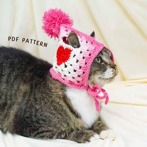 K&ouml;nnte beinhalten: Eine getigerte Katze tr&auml;gt eine handgefertigte rosa, wei&szlig;e und rote H&auml;kelm&uuml;tze mit Herzmuster und einem rosa Bommel. Die M&uuml;tze ist mit einer rosa Schnur unter dem Kinn der Katze gebunden. Der Text "PDF PATTERN" ist sichtbar.