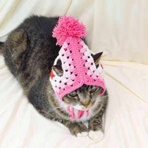 K&ouml;nnte beinhalten: Eine getigerte Katze mit einer geh&auml;kelten rosa-wei&szlig;en M&uuml;tze mit einem gro&szlig;en rosa Bommel. Die M&uuml;tze hat ein quadratisches Muster und rote Sternakzente. Die Katze liegt auf einer wei&szlig;en Oberfl&auml;che.