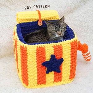 May include: A crocheted cat bed in yellow and orange stripes, with a blue star and a blue interior. A cat is inside the bed. The text "PDF PATTERN" is visible. The bed has a handle and is made of yarn.