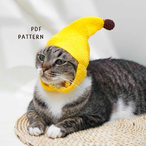 May include: A tabby cat wearing a bright yellow crocheted banana hat and collar. The hat has a brown tip. The text "PDF PATTERN" is visible in the image. The cat is lying on a woven mat.