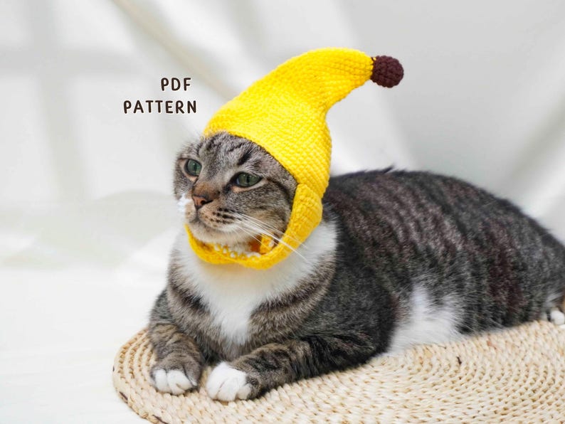 May include: A tabby cat wearing a bright yellow crocheted banana hat and collar. The hat has a brown tip. The text "PDF PATTERN" is visible in the image. The cat is lying on a woven mat.