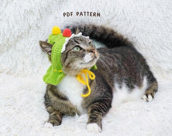 May include: A tabby cat wearing a green crocheted hat with yellow and red accents. The hat is tied with a yellow string. The text "PDF PATTERN" is visible in the background. The cat is lying on a white, fluffy surface.