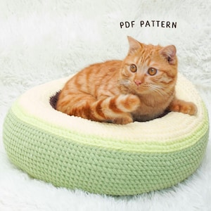 May include: A ginger cat rests in a round, crocheted pet bed. The bed features a light green exterior, a cream-colored interior, and a brown base. The text "PDF PATTERN" is visible in the image.