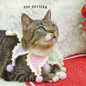 May include: A tabby cat wearing a colorful, crocheted collar with a jester-like design. The collar features pastel pink, green, and lavender sections with small pom-poms. The text "PDF PATTERN" is visible. The cat is on a white fluffy surface.