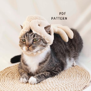 May include: A beige crocheted octopus hat worn by a tabby cat with white markings. The hat features a rounded top with dangling tentacles and the text PDF PATTERN printed in black. The cat is resting on a round woven mat with a neutral background.