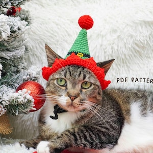 Könnte beinhalten: Eine getigerte Katze mit einem rot-grünen Elfenhut mit rotem Bommel. Die Katze liegt vor einem Weihnachtsbaum mit roter Kugel. Der Text "PDF PATTERN" ist sichtbar.