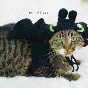 Puede incluir: Un gato atigrado con un disfraz de dragón negro de ganchillo, con alas y un tocado con detalles verdes y blancos. El texto "PDF PATTERN" es visible. El gato está sobre una superficie blanca.