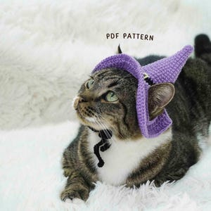 Puede incluir: Un gato atigrado con un sombrero de bruja morado tejido a ganchillo y un collar negro. El gato está tumbado sobre una superficie blanca y esponjosa. El texto "PDF PATTERN" es visible encima del gato.