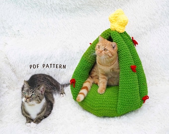 Puede incluir: Una cama para gatos de ganchillo verde con forma de árbol de Navidad, adornada con una estrella amarilla y detalles rojos. Un gato atigrado naranja está dentro de la cama, mientras que un gato atigrado gris está cerca. El texto "PDF PATTERN" es visible.