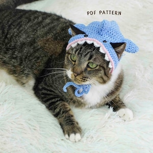 Könnte beinhalten: Eine getigerte Katze trägt einen hellblauen gehäkelten Haifischhut mit rosa und weißen Zähnen und schwarzen Augen. Die Katze liegt auf einer weißen, flauschigen Oberfläche. Der Text "PDF PATTERN" steht über der Katze.