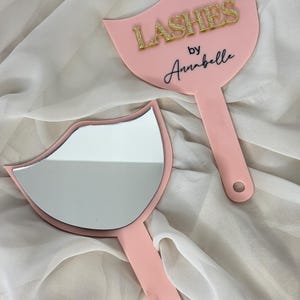 Op de afbeelding: Twee roze handspiegels met een lipvormig ontwerp. Eén spiegel heeft een reflecterend oppervlak, de andere heeft de tekst "LASHES by Annabelle" in gouden glitter en zwart schrift. De spiegels hebben een handvat met een gat om op te hangen.