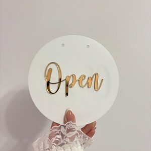 Op de afbeelding: Een witte, ronde plaat met het woord "Open" in gouden letters. De plaat heeft twee kleine gaatjes aan de bovenkant en wordt vastgehouden door een persoon met een witte kanten mouw.