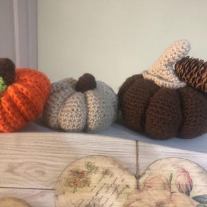 May include: Three crocheted pumpkins, one orange, one brown, and one beige. The brown pumpkin has a small pine cone on top.