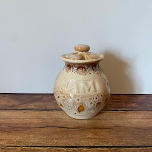 May include: A ceramic jar with a lid, featuring a beige base with brown speckles and a brown rim. The word "JAM" is embossed on the front. The jar sits on a wooden surface.