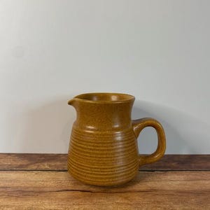 May include: A brown ceramic jug with a textured surface and a handle. The jug is sitting on a wooden surface.