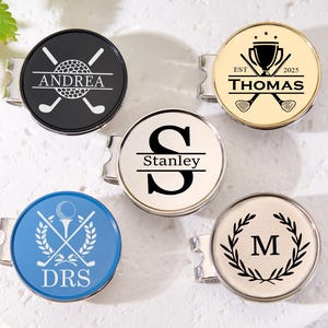 May include: A collection of personalized golf ball markers. Each marker features a circular design with various graphics, including golf balls, crossed clubs, initials, and names. Colors include black, white, gold, and blue. The markers have a silver clip.