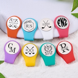Könnte beinhalten: Eine Sammlung personalisierter Golfballmarker in verschiedenen Farben, darunter Orange, Blau, Weiß, Rosa, Lila, Gelb und Türkis. Jeder Marker hat ein einzigartiges Design mit Initialen, Namen oder Logos und Golfschläger-Grafiken.