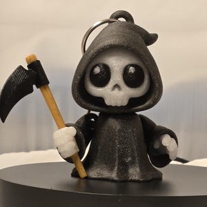 May include: A black Grim Reaper figurine with a scythe, wearing a hooded cloak. The figurine has a skull face with large, round eyes and white gloves. The scythe has a wooden handle and a black blade. The figurine is on a black platform.