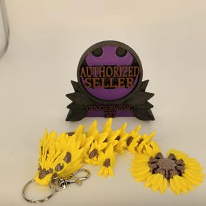 May include: A yellow dragon keychain with a silver clasp and ring, next to a yellow sunflower and a purple and black "AUTHORIZED SELLER" emblem. The dragon has brown accents and a detailed design.