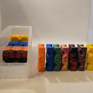 May include: A collection of colorful, geometric fidget toys in various colors, including orange, blue, green, pink, and yellow. Some are stored in a clear plastic container, while others stand upright.