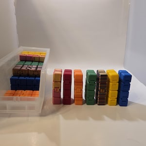 May include: A collection of colorful fidget toys in various colors, including pink, red, orange, green, brown, yellow, and blue. Some are stacked in a clear plastic bin, while others are arranged in a row.