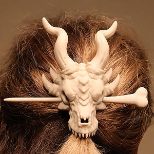 May include: Ivory-colored hair accessory featuring a dragon skull design with horns and a bone-shaped pin. The skull has detailed features, including teeth and eye sockets. The accessory is worn in brown hair.