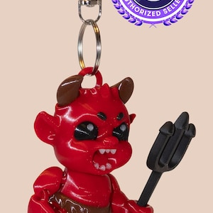 May include: A red devil keychain with brown horns and a pitchfork. The devil figure has a cartoonish design with a wide grin and black eyes. The keychain includes a silver clasp and ring. The Printverse Authorized Seller logo is in the background.