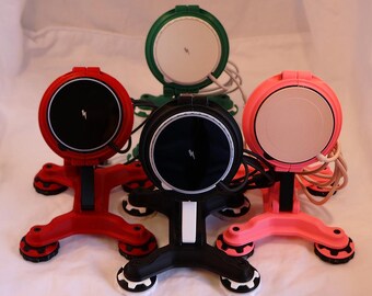 May include: A collection of wireless chargers in red, black, green, and pink. Each charger has a circular charging surface with a lightning bolt symbol and a stand with suction cup feet. Cables are attached to each charger.