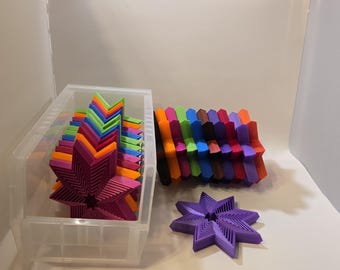 May include: A collection of colorful, star-shaped objects in a clear plastic bin and arranged on a white surface. The stars are various colors, including purple, orange, green, and blue. One star is isolated on the surface.