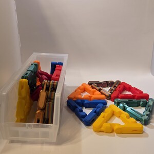 May include: A clear plastic bin filled with colorful, interlocking plastic shapes. Several assembled shapes in various colors, including red, blue, yellow, and orange, are arranged on a white surface. The shapes appear to be for a construction toy.