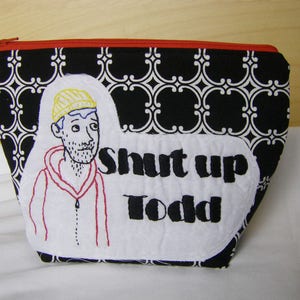 May include: Black and white patterned fabric zippered pouch with a white embroidered design of a person wearing a yellow hat and the text "Shut up Todd".