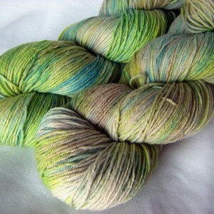 Op de afbeelding: Een bol garen in tinten van groen, blauw en beige. Het garen is handgeverfd en heeft een gevarieerd kleurpatroon.