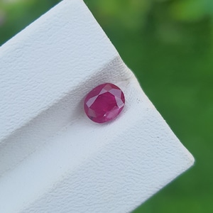May include: A faceted, oval-shaped ruby gemstone. The gem is a deep red color and sits on a white, textured surface. The background is a blurred green.