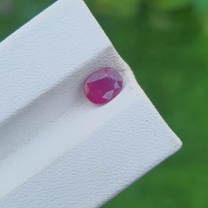 Op de afbeelding: Een gefacetteerde, ovaalvormige robijn edelsteen, met een dieprode kleur. De edelsteen ligt op een wit, getextureerd oppervlak, met een vage groene achtergrond. De facetten van de robijn vangen het licht.