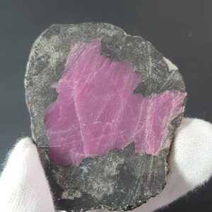 Op de afbeelding: Een ruw geslepen edelsteen met een levendig roze centrum en een donkergrijze buitenlaag. De steen heeft een natuurlijke, onregelmatige vorm, die de ruwe, ongepolijste textuur laat zien. Het roze gebied heeft een kristallijn uiterlijk.