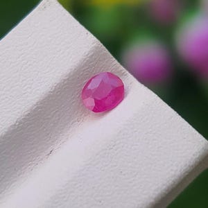 May include: A faceted, oval ruby gemstone, with a vivid pink-red colour. The gem is set against a white, textured surface, with a blurred green and pink background. This image showcases a precious stone.