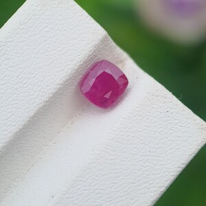 May include: A cushion-cut ruby gemstone, displaying a deep red hue. The gemstone is set against a textured white surface, with a blurred green background. The ruby's facets catch the light, showcasing its brilliance.