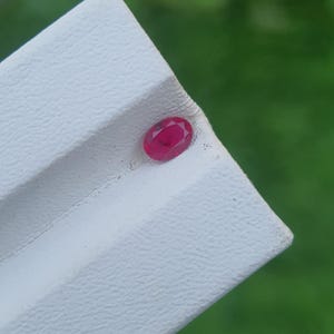 May include: A small, oval-cut ruby gemstone, displaying a rich red hue. The gem is set on a white, textured surface. The background is a blurred green, suggesting an outdoor setting.